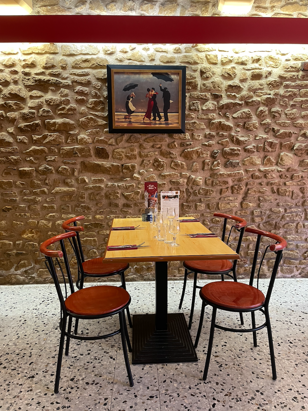 Table à l'intérieur du restaurant dressée pour le repas du midi avec mur en pierres dorées et tableau représentant un couple qui danse